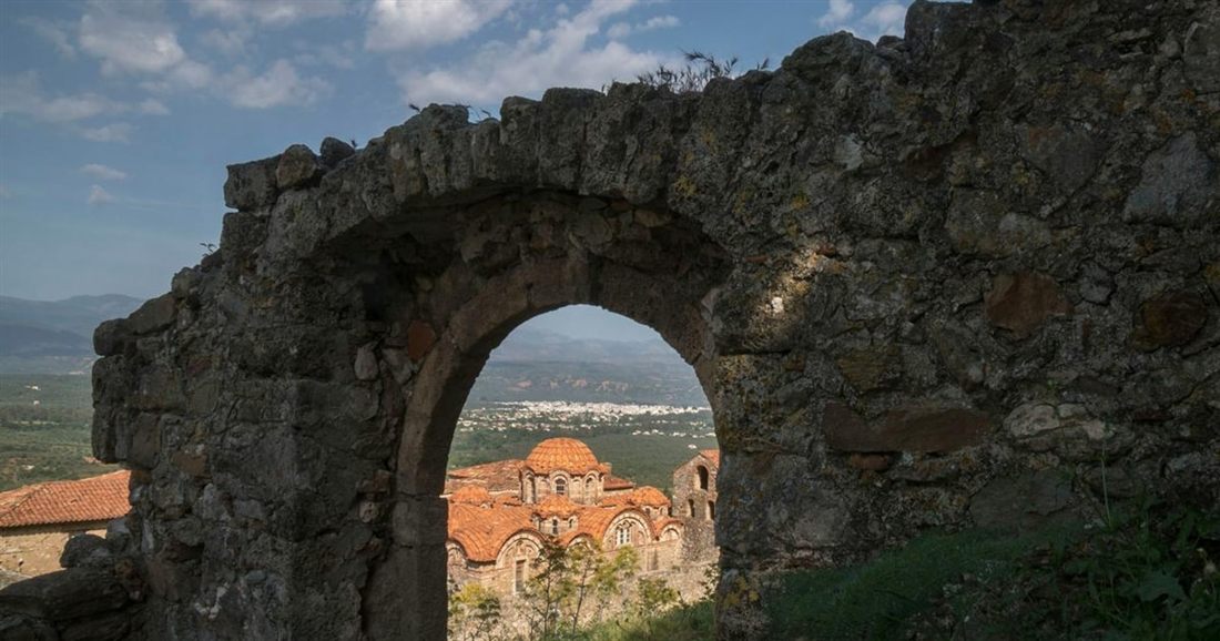 A stone arch in a ruined wall at Mystras frames a Byzantine church with red-tiled domes on the slope below.