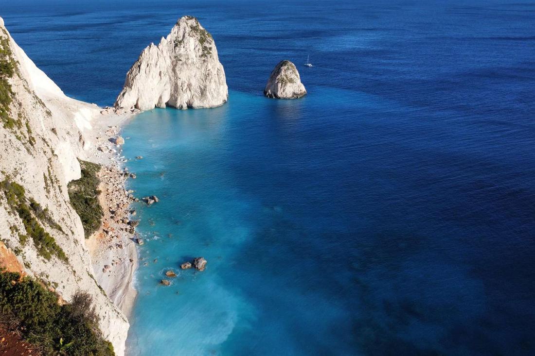 White Myzithres rocks rising from deep blue sea beside a white cliff and pebble cove, seen from the viewpoint above Keri