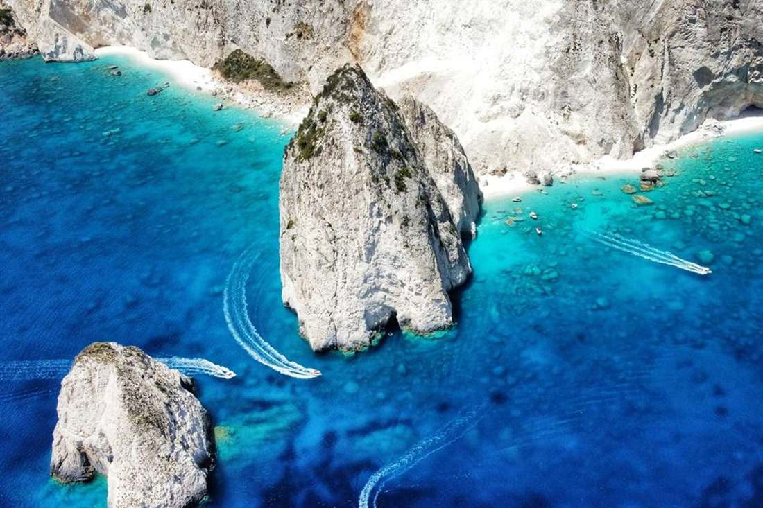 Aerial view of the two white Myzithres rocks and turquoise bay with speedboat wakes beneath the white cliffs