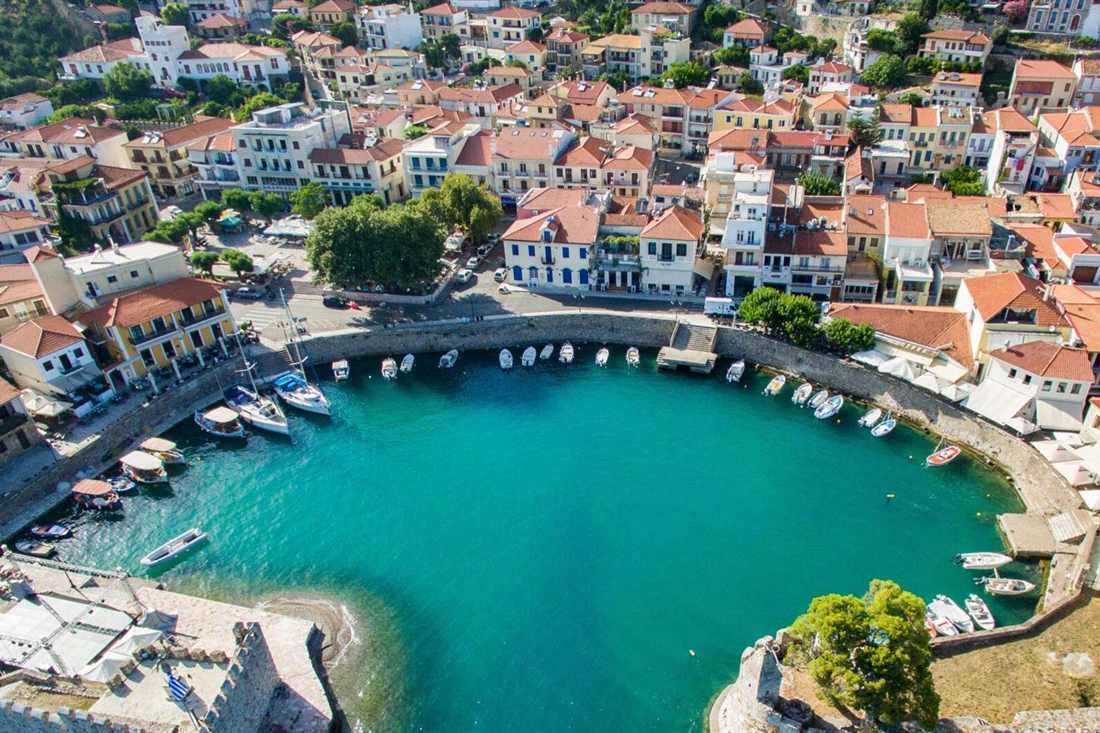 Aerial view of the round harbour of Nafpaktos with moored boats along the stone quay, red-roofed houses behind and turquoise water in front.