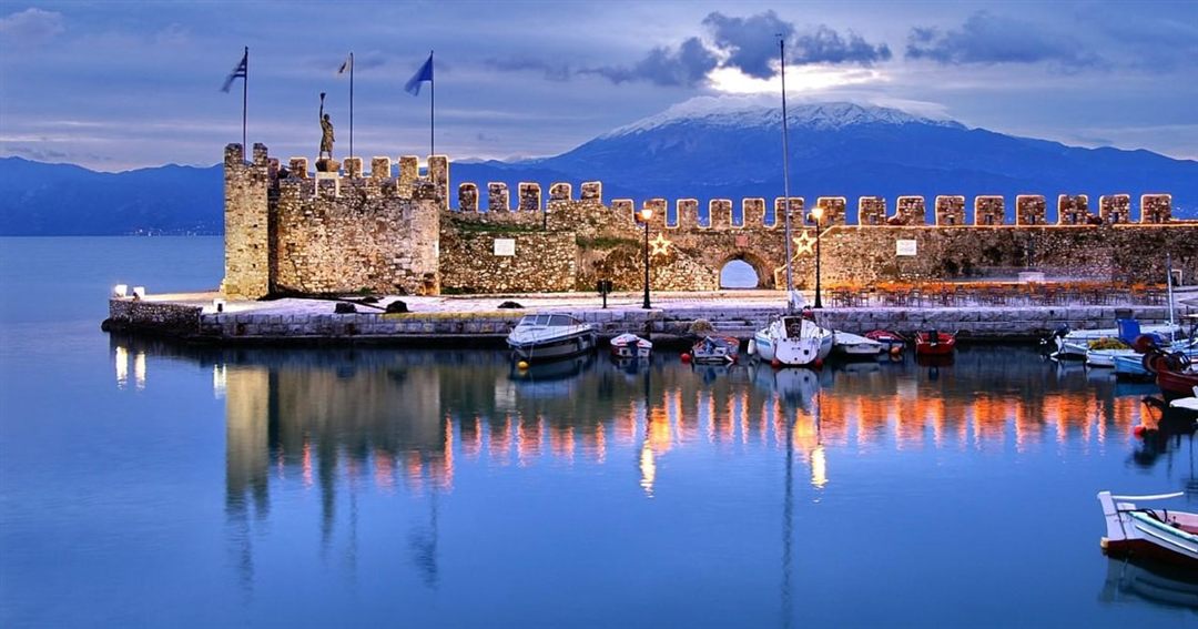 The stone tower and battlements at the entrance of the Venetian harbour of Nafpaktos at dusk, with snow-capped mountains across the gulf.