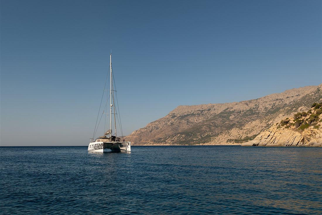 A sailing catamaran at anchor in calm blue water off a rocky Greek coast, the type of boat used on a Nafplio catamaran cruise.