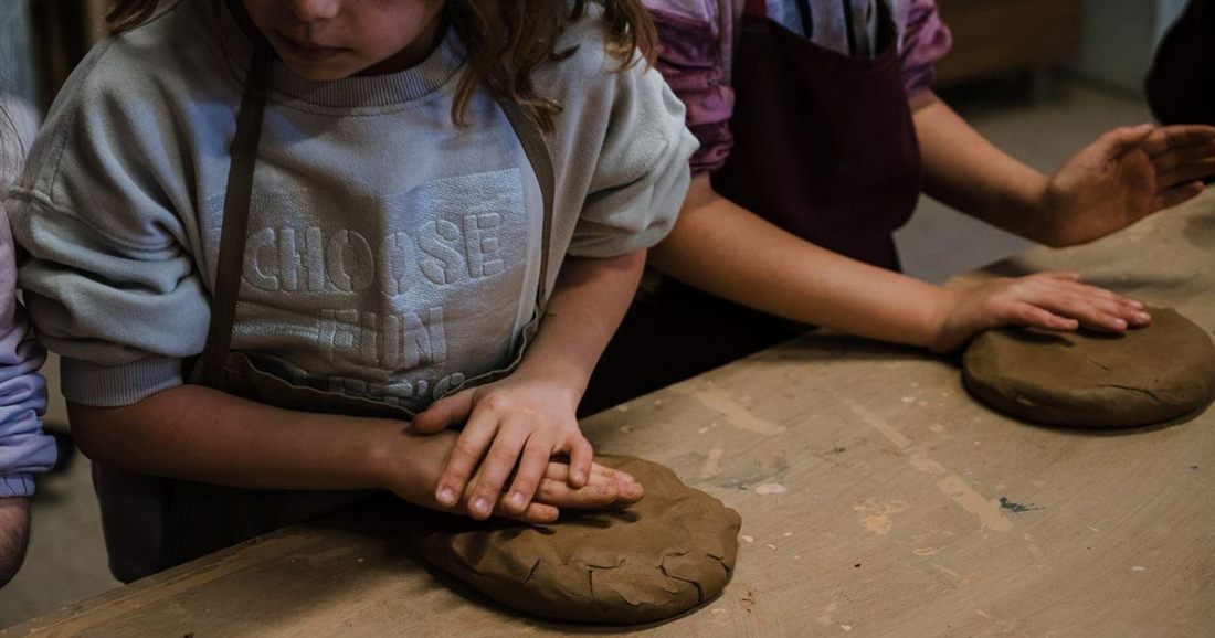 A child presses a disc of brown clay flat on a wooden workbench beside other hands at work, in a Nafplio pottery class.