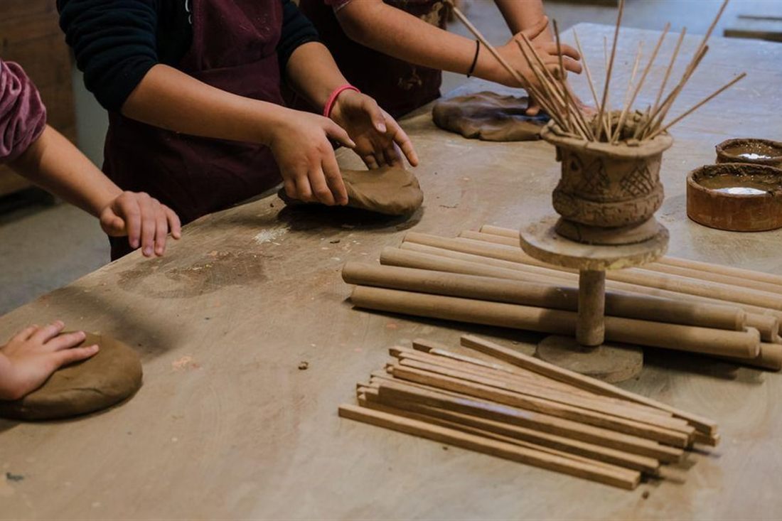 Hands press discs of clay on a wooden workbench beside a coiled clay pot and bundles of wooden sticks, in a Nafplio pottery class.