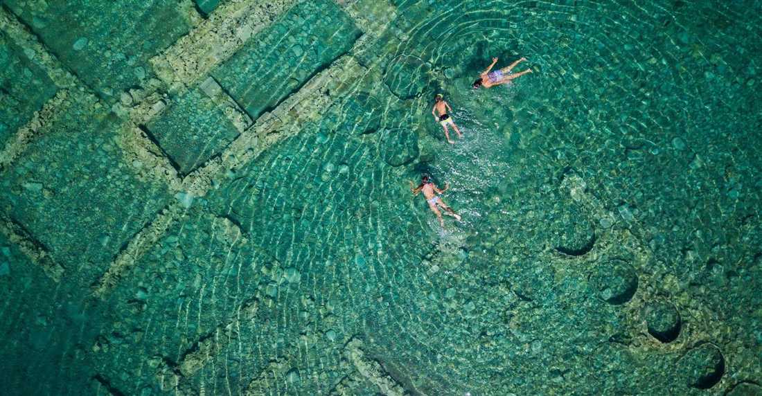 Snorkellers swimming over the stone foundations of the sunken city in clear green water near Old Epidaurus