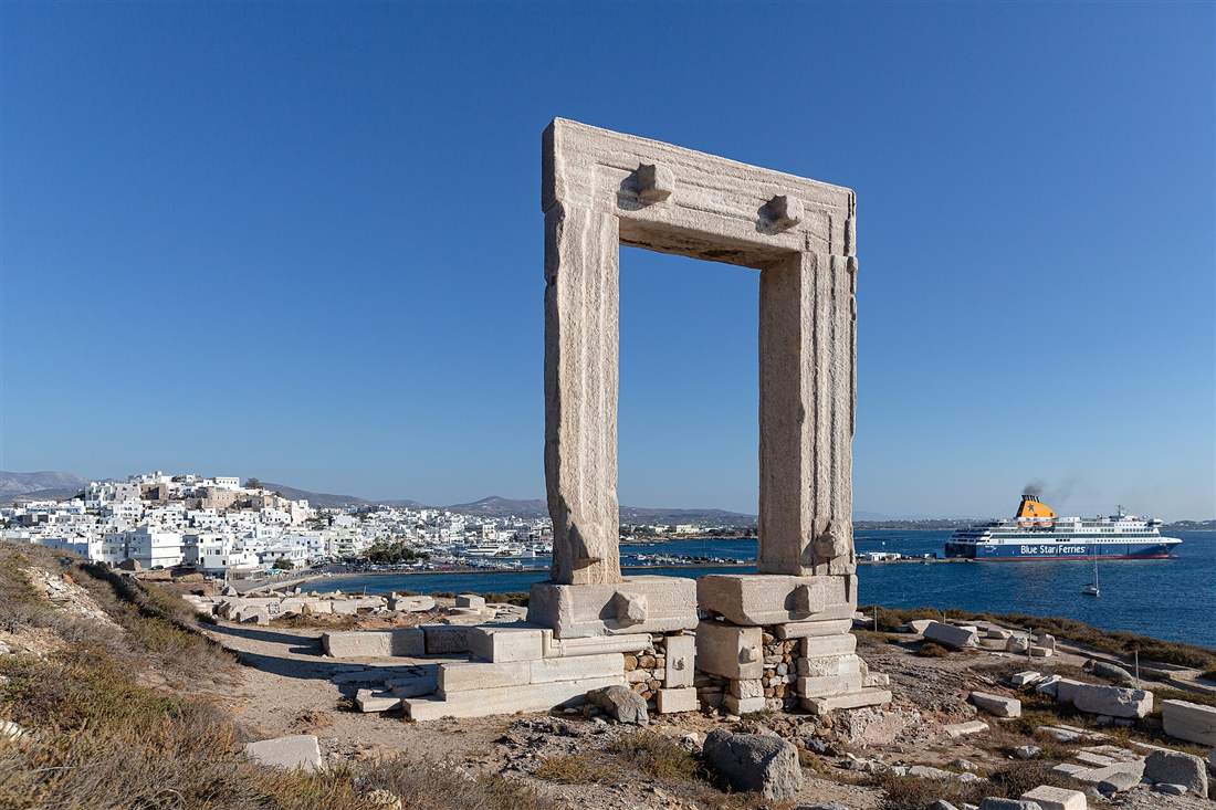 The marble Portara gateway of the Temple of Apollo on Naxos, with Chora and a ferry in the harbour behind under a clear sky.