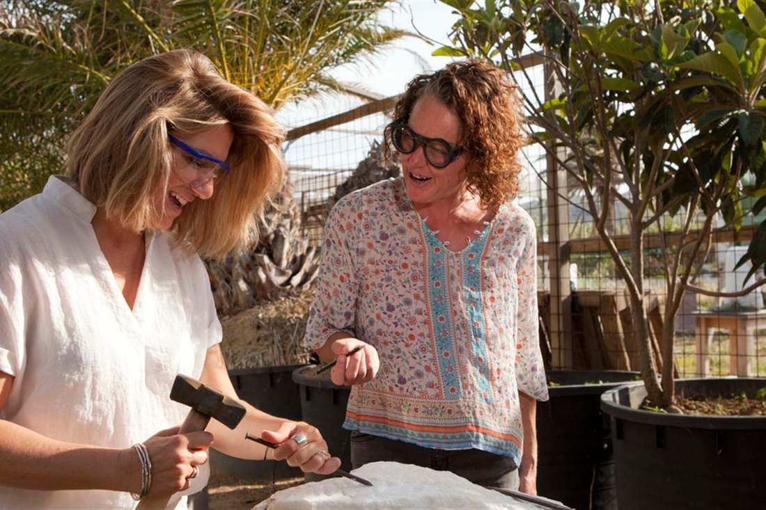 Two women in safety glasses work a marble block with a hammer and chisel in a garden studio during Naxos marble carving.