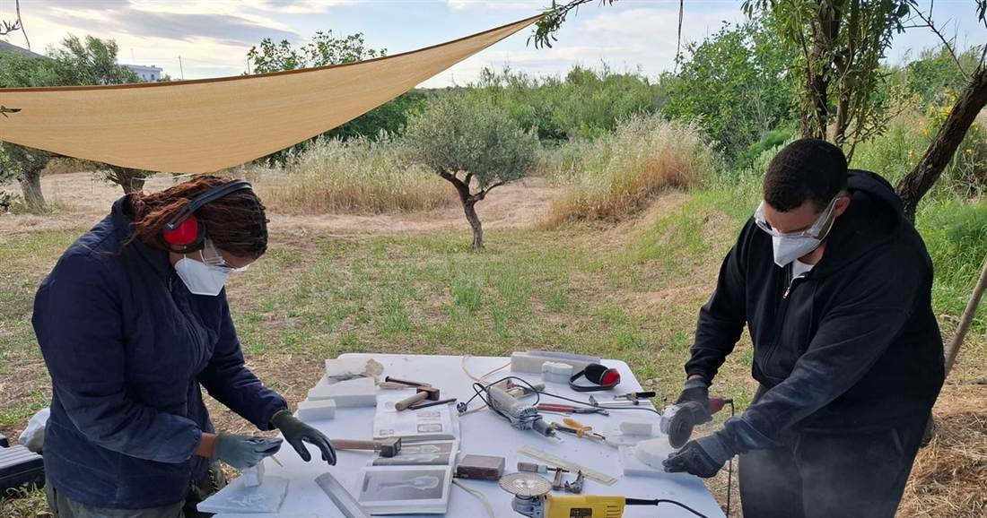 Two people in masks and gloves file small pieces of white marble at a tool-covered table under a shade sail in an olive grove during Naxos marble carving.