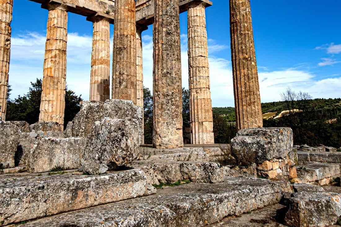 Tall limestone columns of the Temple of Zeus at Ancient Nemea standing above fallen blocks under a blue sky.