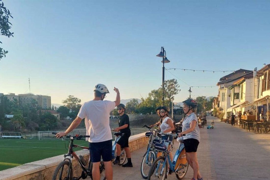 Cyclists in helmets with bicycles stop on a stone promenade above a green football pitch, with cafe tables and old houses along the street