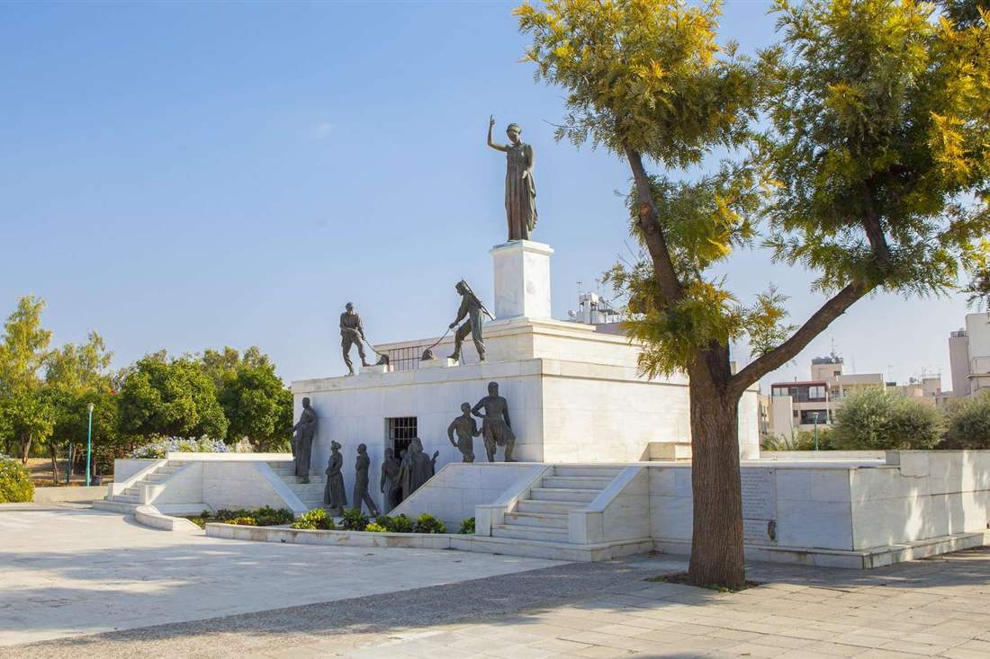 White marble Liberty Monument in Nicosia with bronze figures and a statue of a woman raising her arm, under a clear blue sky