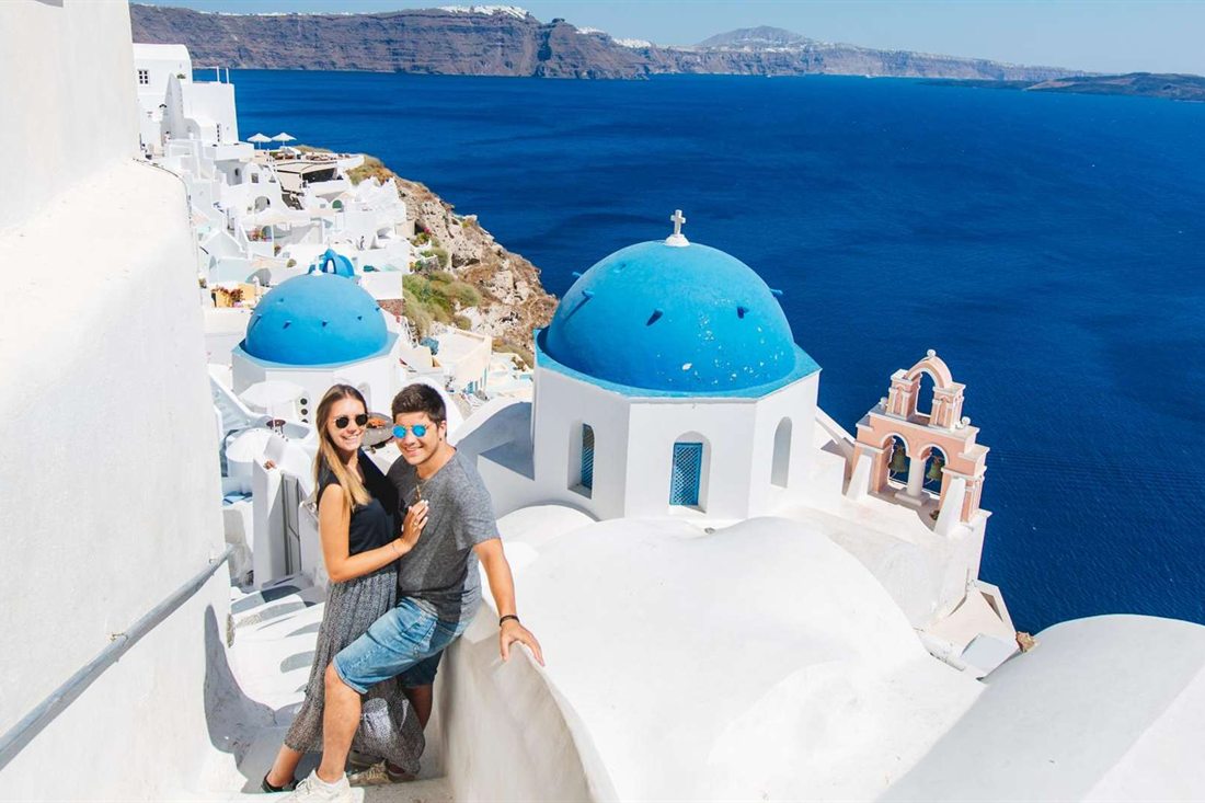 A couple posing together on a whitewashed stairway in Oia with a blue-domed church, a pink bell tower and the deep blue caldera behind them