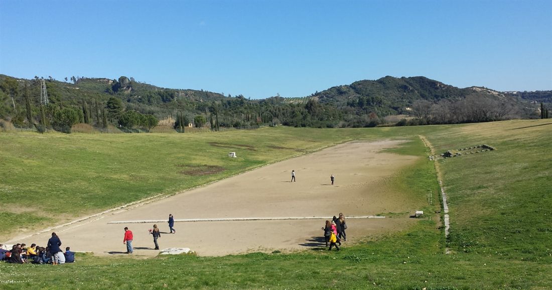 The long earth track of the ancient stadium at Olympia between grassy banks, with visitors walking on it, a stop on an Olympia day trip from Kalamata.