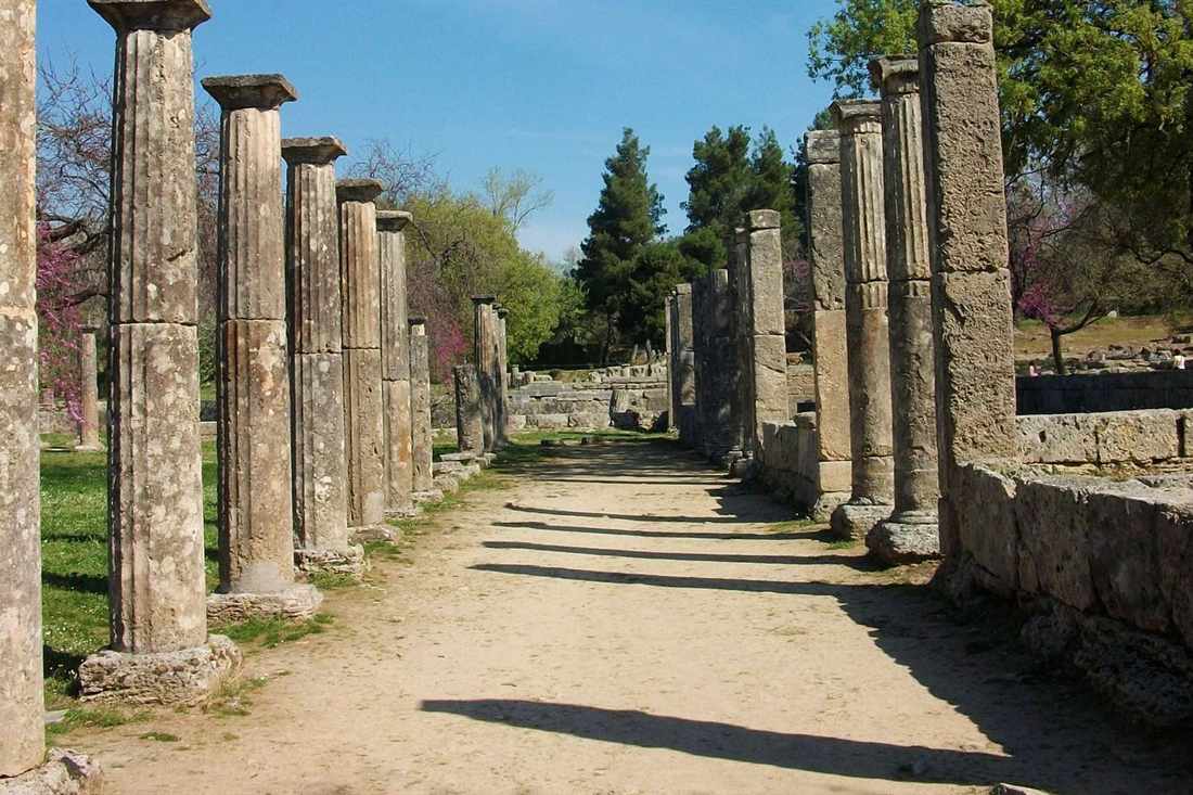 A row of Doric columns along a gravel path in the ruins of Ancient Olympia under a blue sky