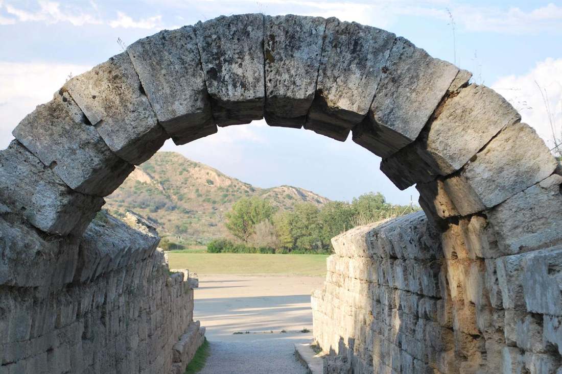 The stone arch of the vaulted entrance to the ancient stadium at Olympia, with the running track beyond