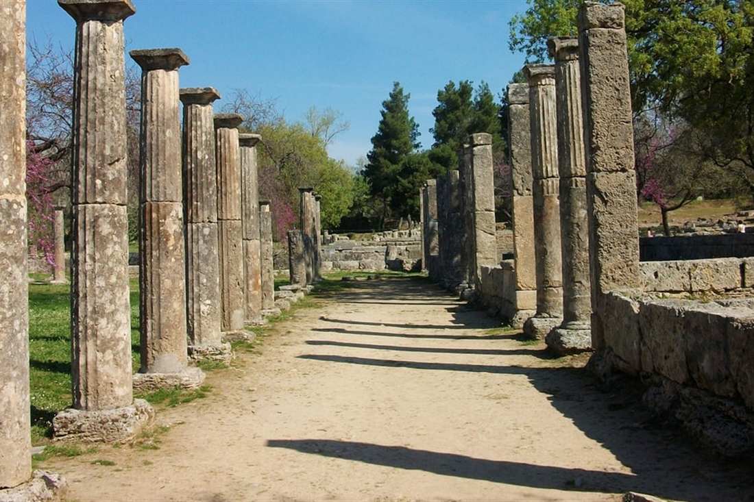 A row of standing stone columns along a sunny path at Ancient Olympia, with pine trees and blue sky.