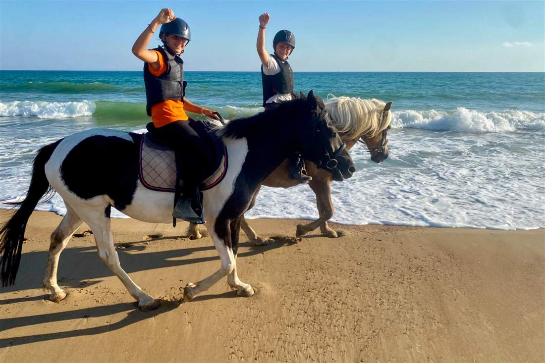 Two young riders in helmets on horseback along a sandy beach as waves break beside them