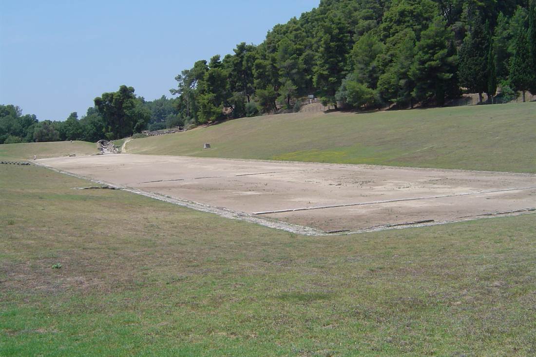 The long earth running track of the ancient stadium at Olympia, with grassy banks and pine trees around it