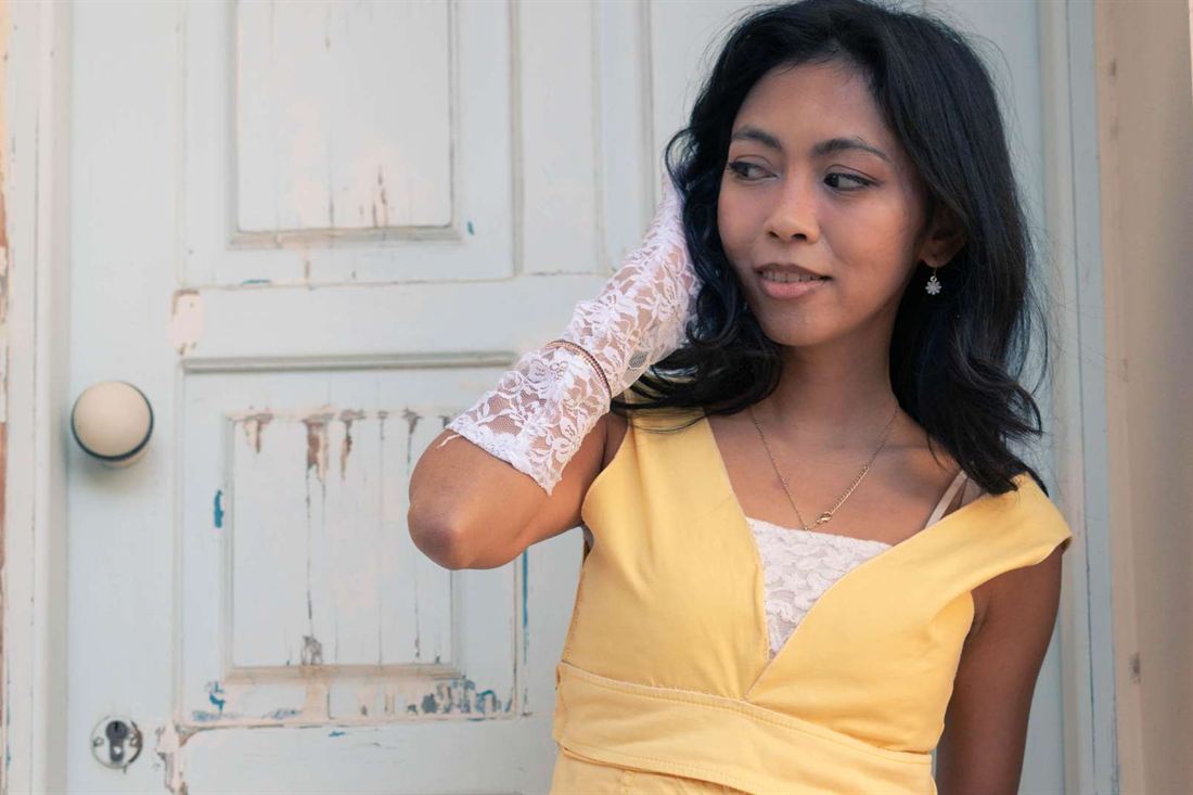 Woman in a yellow dress and white lace glove posing for the camera against a weathered white wooden door