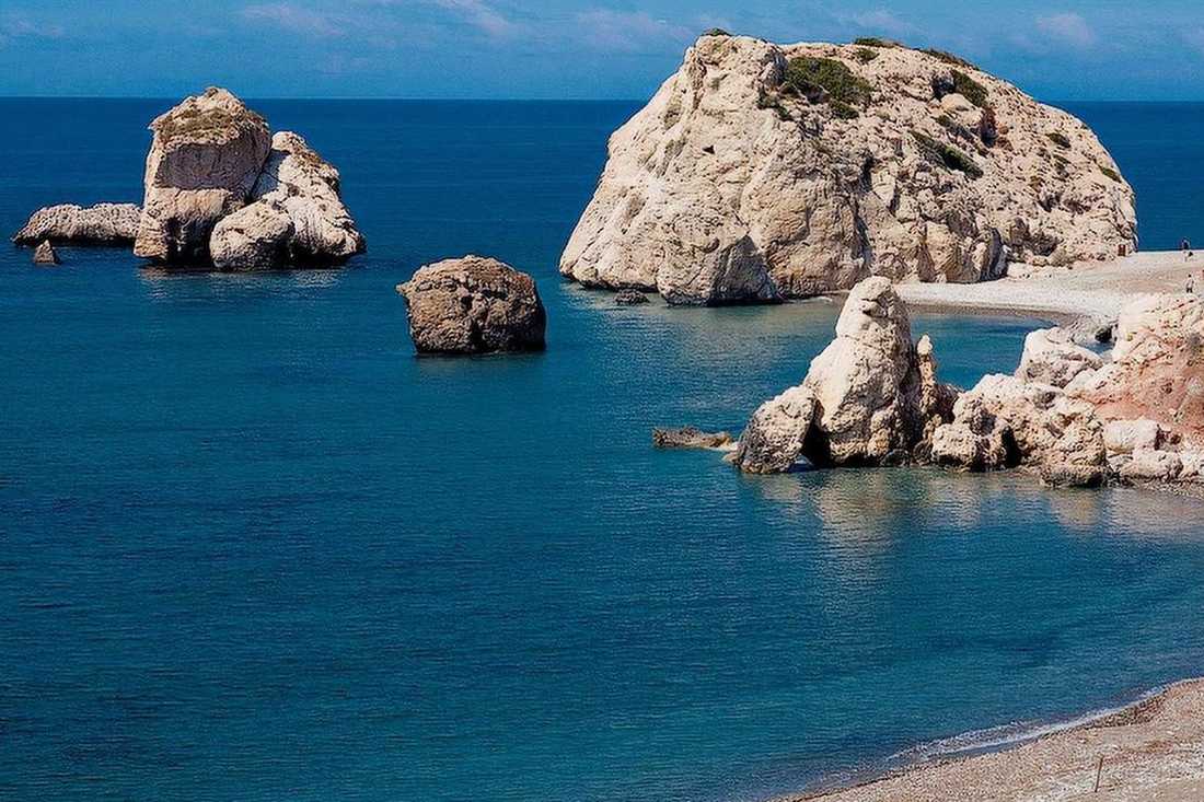 The rock and sea stacks of Petra tou Romiou rising from blue water beside a pebble beach