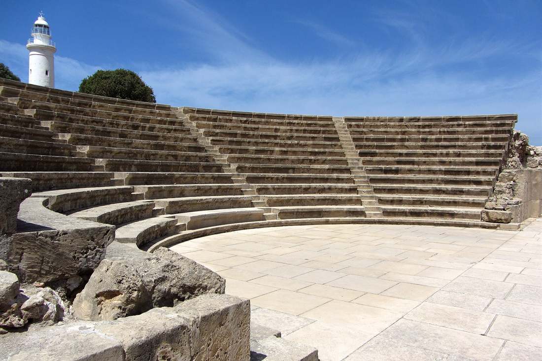 Curved stone seating of the ancient odeon in Paphos Archaeological Park with a white lighthouse and green tree behind