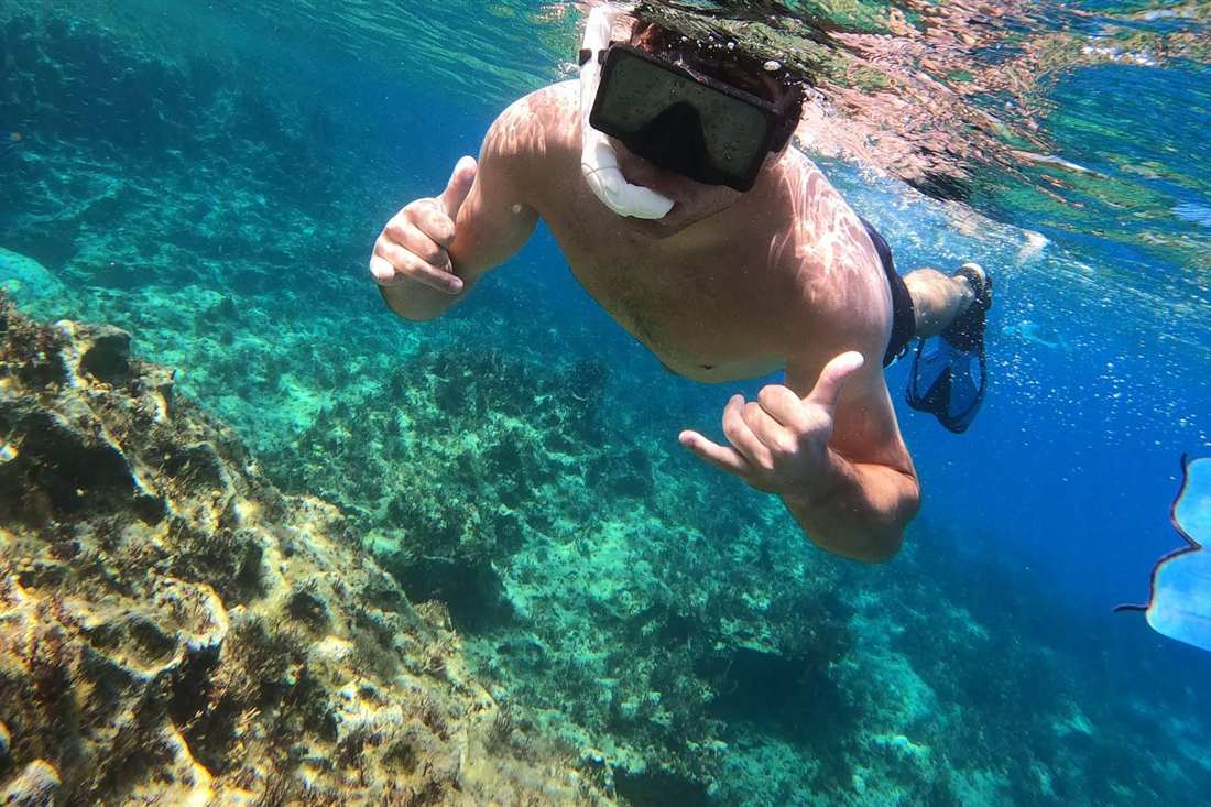 A snorkeler with a mask, snorkel and fins gives a thumbs up above a rocky reef in clear blue water