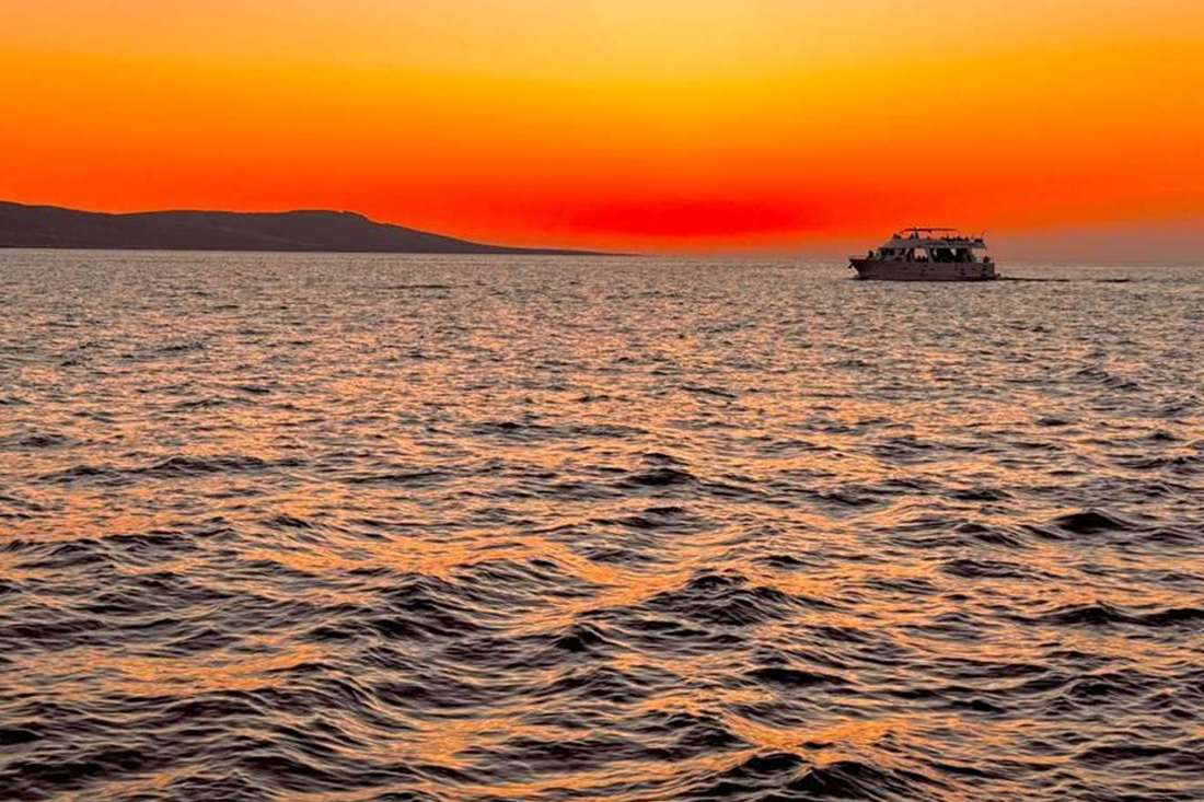 A cruise boat sailing on the sea at sunset under an orange sky with a dark headland on the left