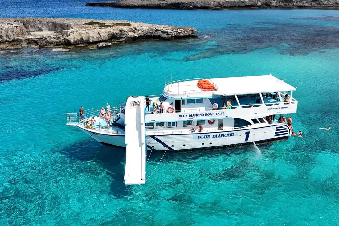 A white two-deck Blue Diamond cruise boat with a white slide moored in turquoise water beside a rocky shore