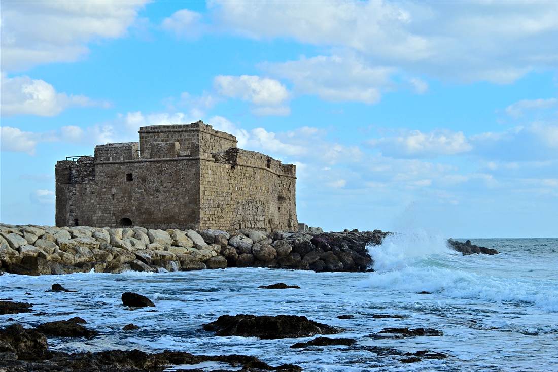 Paphos Castle, a square stone fort on the rocky harbour shore with a wave breaking against the breakwater under a blue sky