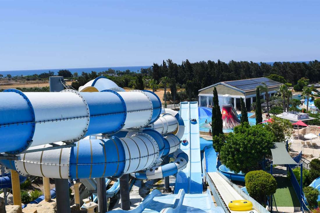 Blue and white tube water slides and a straight racer slide at a water park, with pools, a red volcano feature and sun umbrellas below and the sea on the horizon