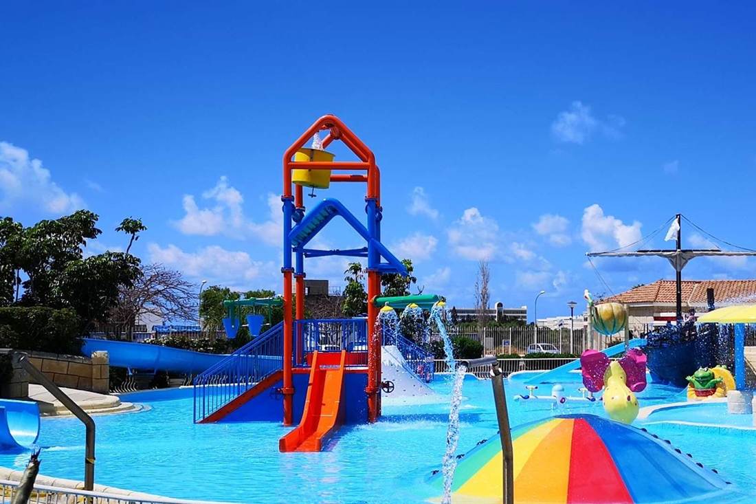 Splash pool for children with an orange and blue water play tower, slides, colourful water toys and a rainbow-striped mushroom fountain under a blue sky