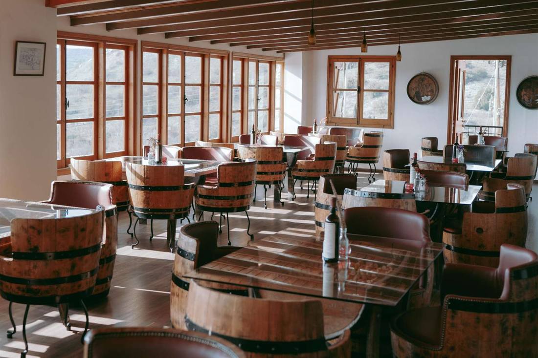 Winery tasting room with barrel-shaped chairs and glass-topped barrel tables holding wine bottles, with tall wooden windows facing a sunlit dry hillside