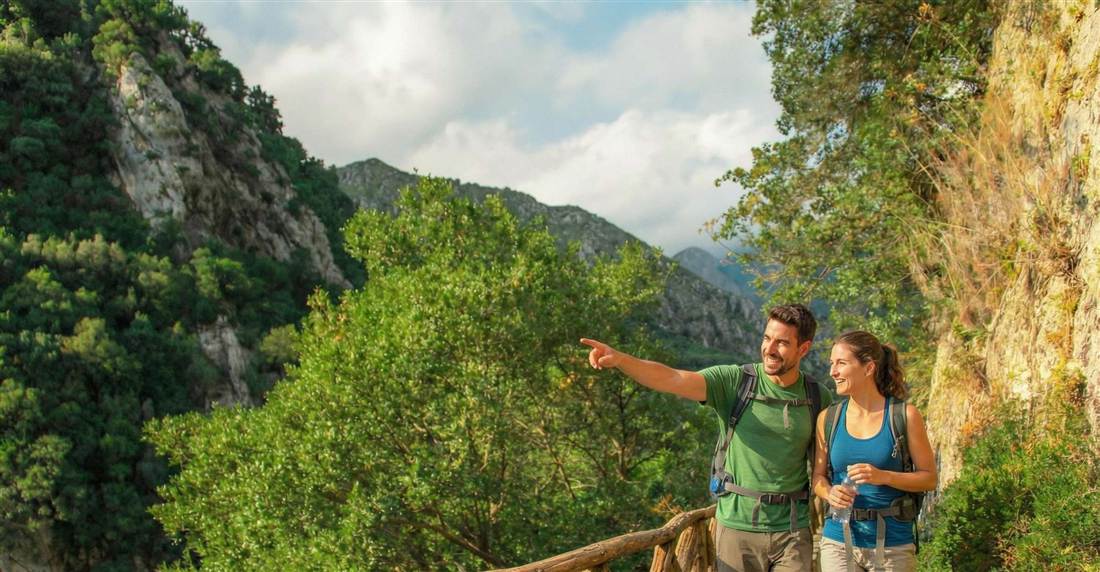 Two hikers with backpacks pointing at the view from a green gorge trail under steep cliffs