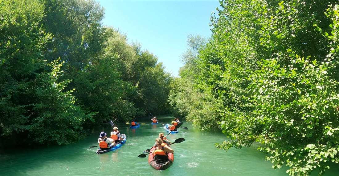 Kayakers in orange life jackets paddling the green Acheron River near Parga between dense trees.