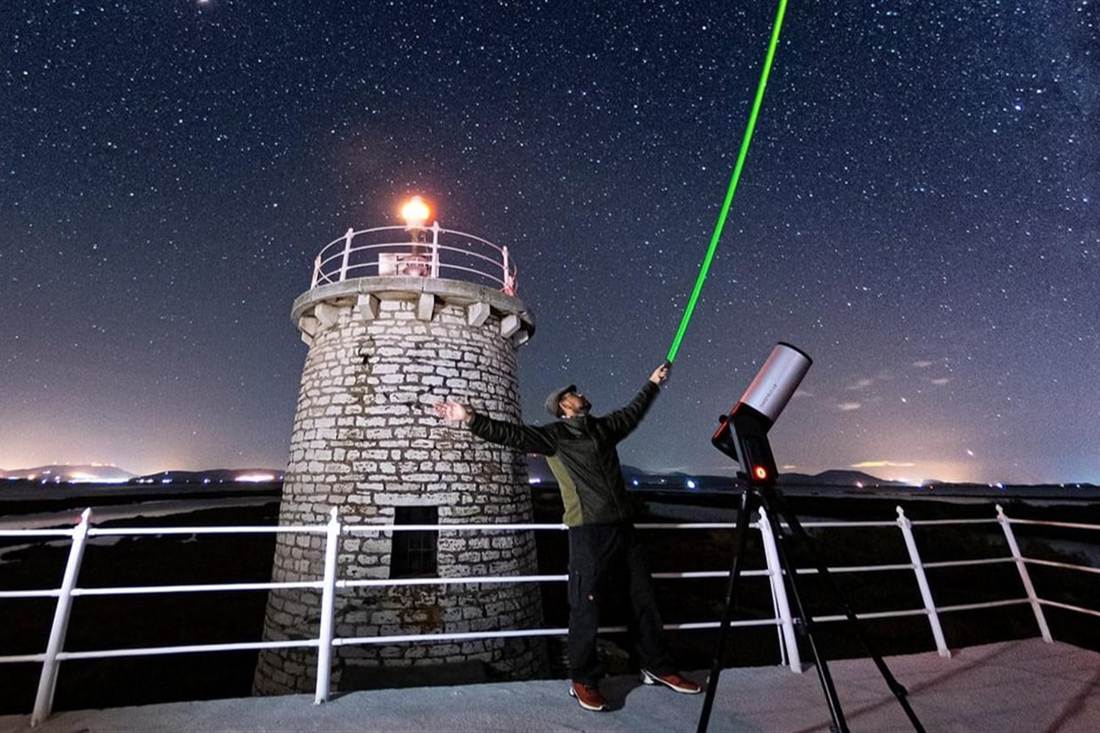 An astronomer pointing a green laser at the stars beside a stone lighthouse and a telescope on a Parga stargazing tour.