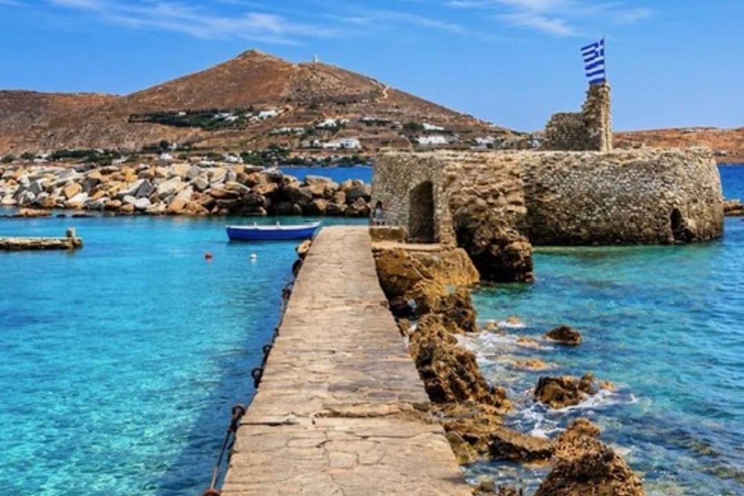 The Venetian fortress at the entrance of Naoussa harbour on Paros, with a stone jetty, clear blue water and a Greek flag on the tower.