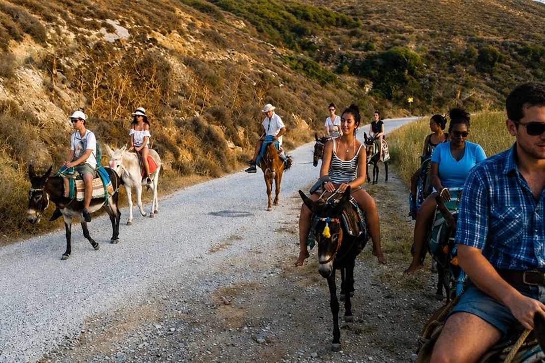 A line of riders on donkeys following a dirt track through dry hills above Parikia on a Paros donkey ride.
