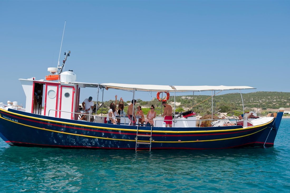 A blue and white traditional wooden fishing boat with guests on deck, anchored in clear water on a Paros fishing trip.