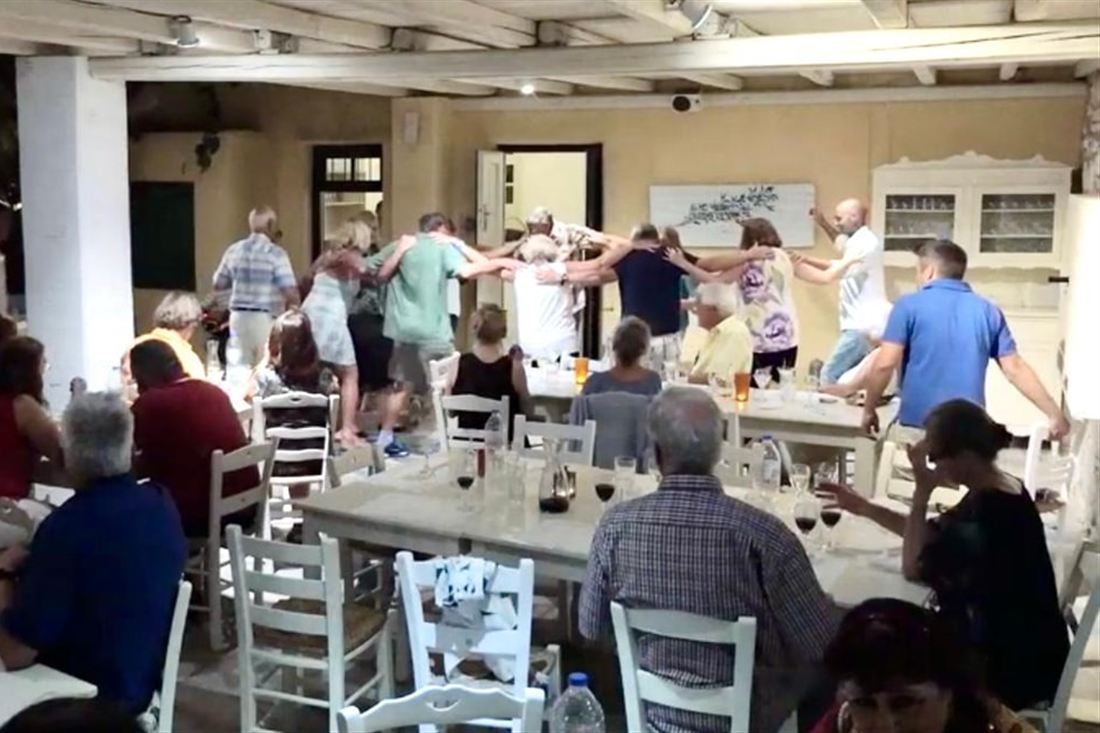 Guests and dancers holding hands in a circle dance between the dinner tables of a taverna at a Paros Greek night.