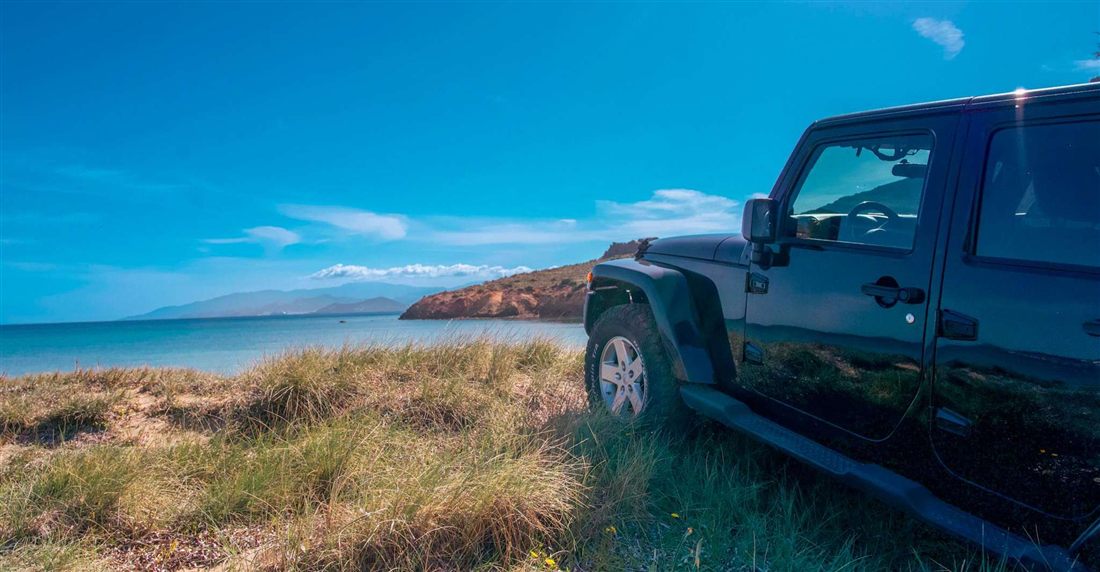 A black jeep parked on a grassy dune above a turquoise bay on the coast of Paros