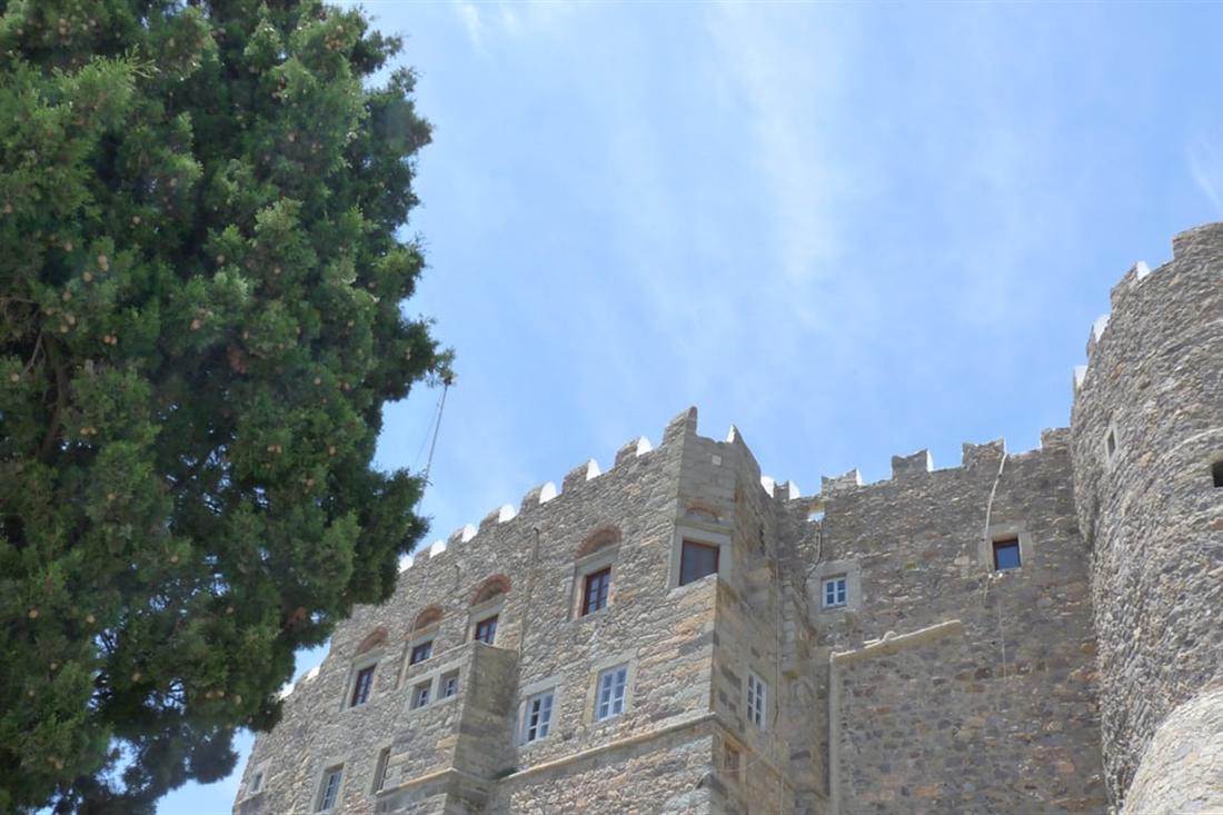 The fortified stone walls and battlements of the Monastery of Saint John on Patmos under a blue sky, beside a tall cypress tree.