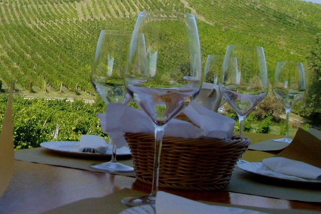 Five empty wine glasses on a table with a bread basket and plates, with terraced vineyard rows on the hillside behind