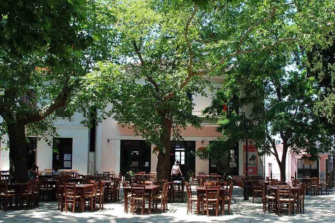 Wooden cafe tables and chairs in a village square shaded by large plane trees in front of a white building