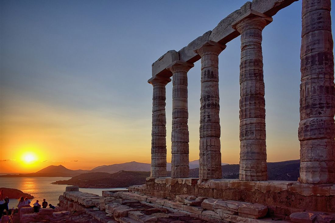 The marble columns of the Temple of Poseidon at Cape Sounion at sunset, with the sun setting over the Aegean Sea and the hills of Attica.