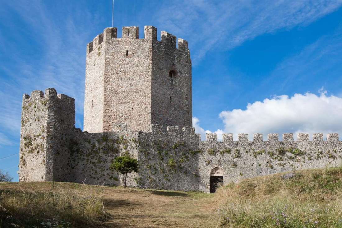 Stone keep and battlemented walls of Platamon Castle on a grassy hill under a blue sky with white clouds
