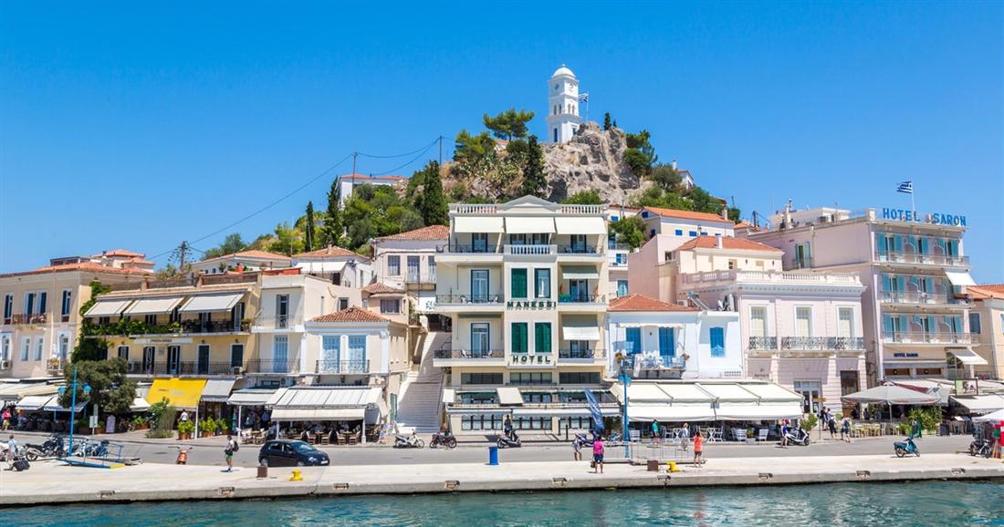 The white clock tower of Poros on a green hilltop above pastel waterfront houses and the harbour quay.