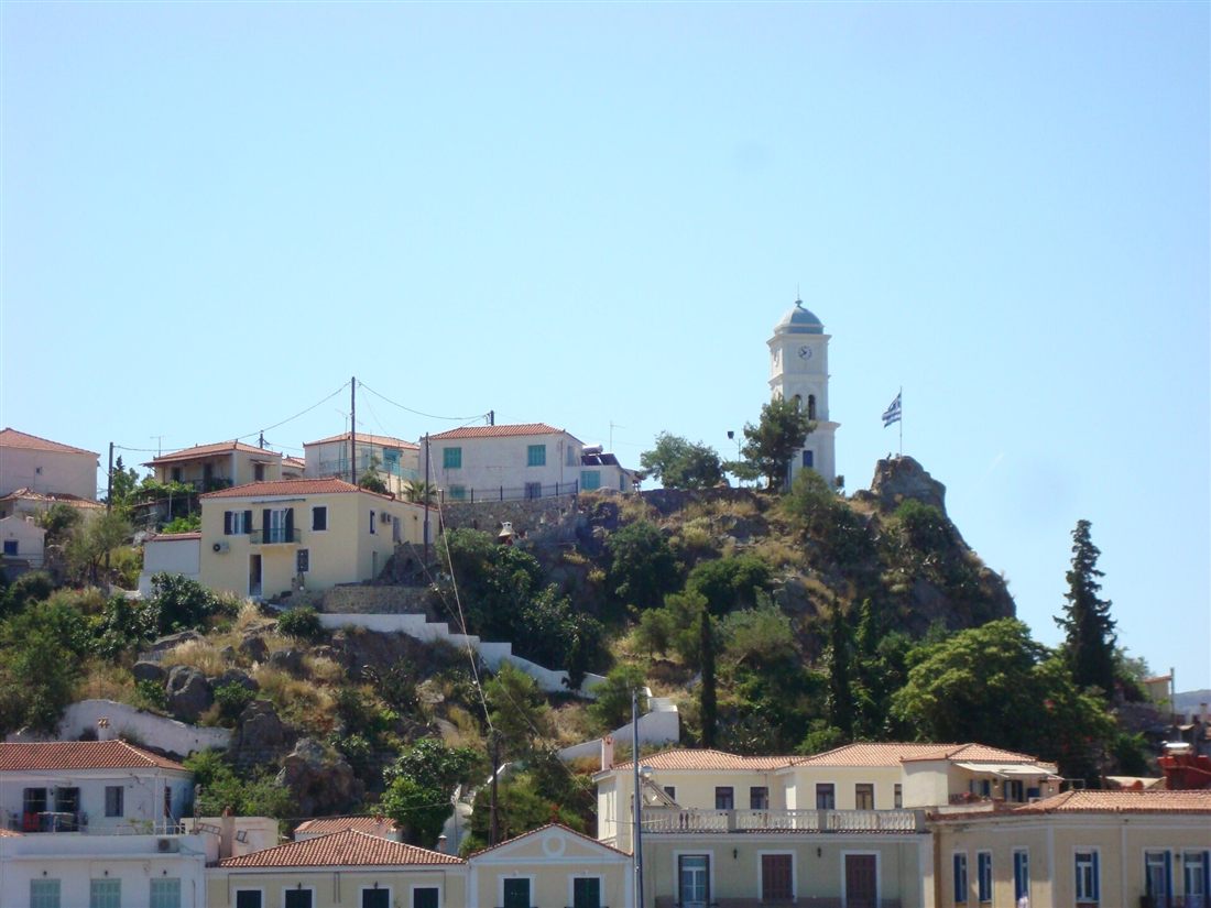 The clock tower of Poros on its rocky hill above the houses of Poros Town under a clear sky