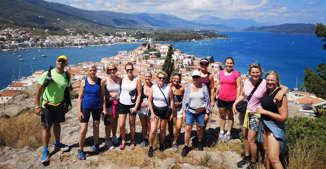 A group of hikers on a hilltop above Poros Town, with the harbour, the strait and the Peloponnese mountains behind them
