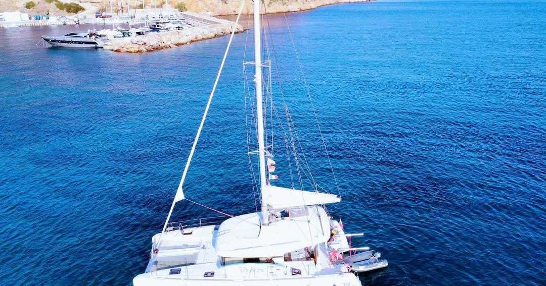 A white sailing catamaran on deep blue water off the coast, seen from above.