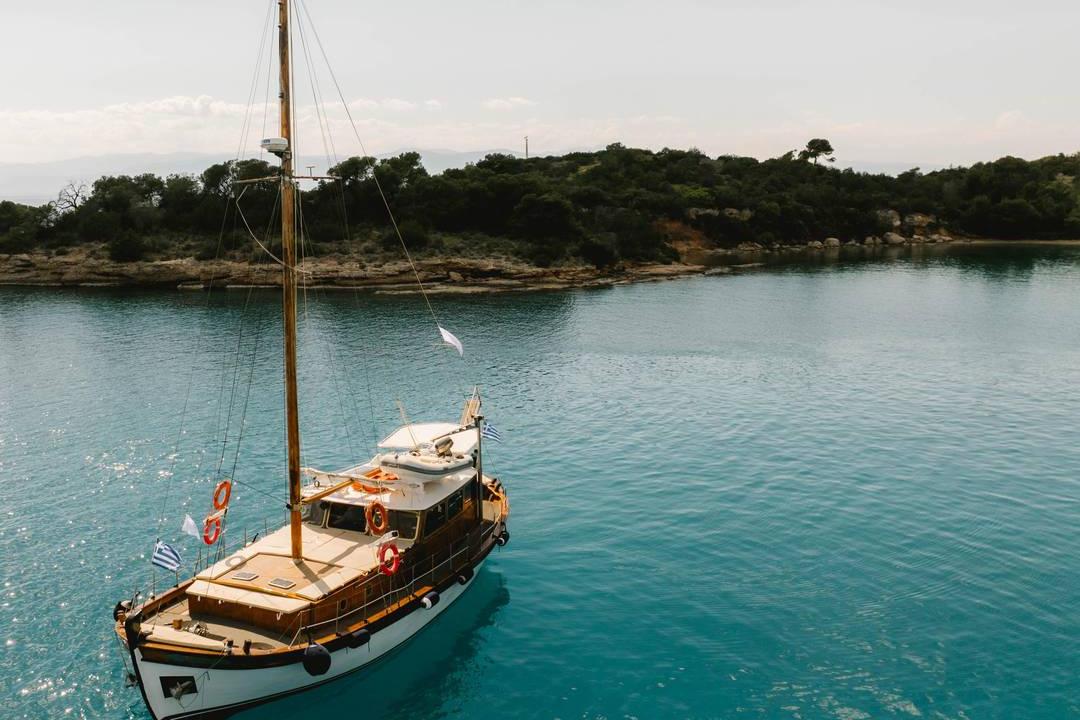 A traditional wooden sailing boat anchored in a turquoise bay below a pine-covered shore near Porto Heli.